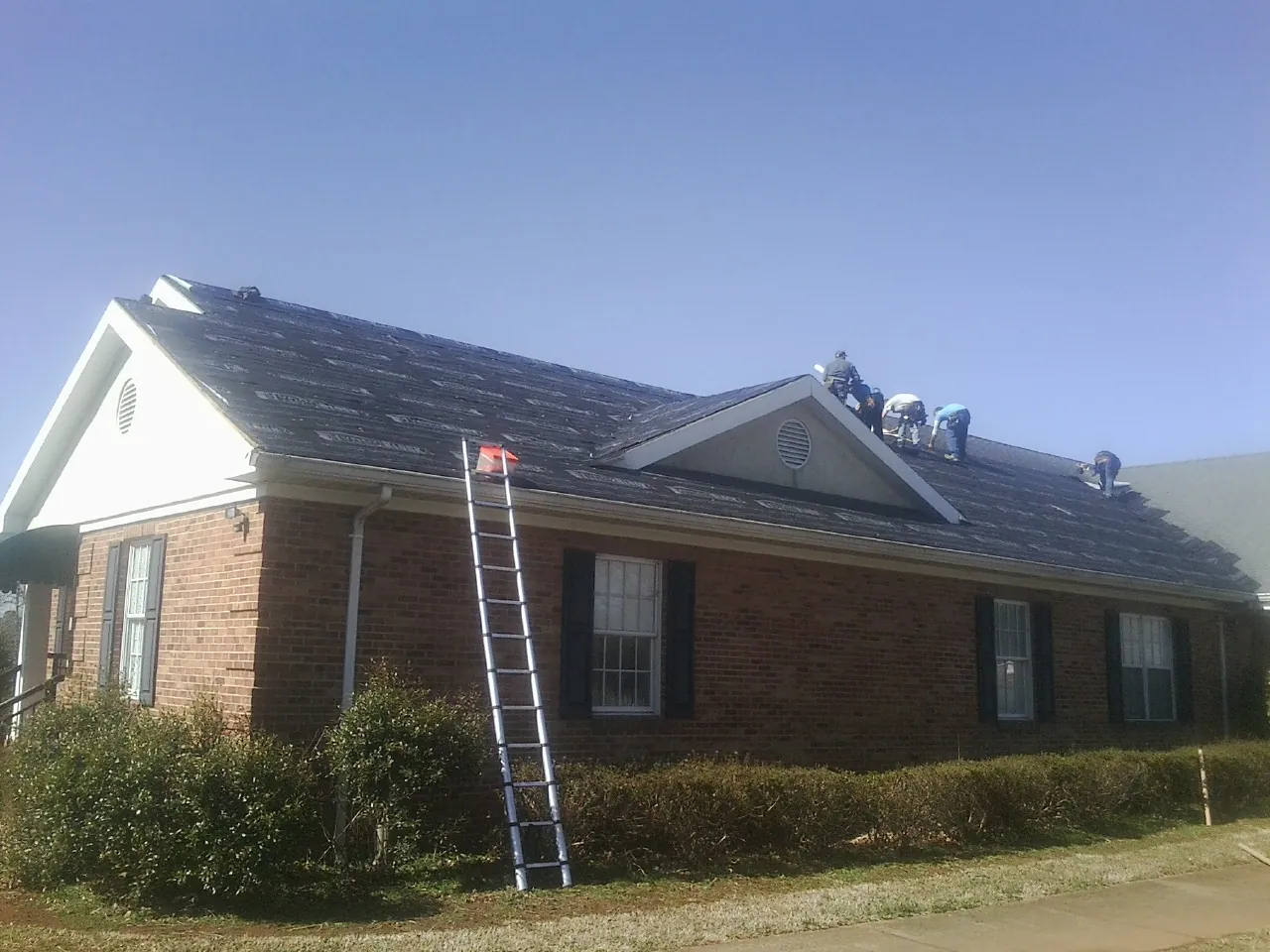 Crew tearing shingles off a brick ranch roof with a ladder set — Metal Roof Repair work by our crew in Searsboro