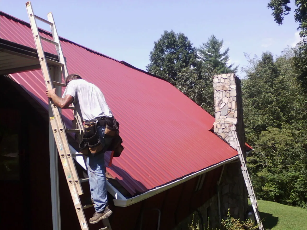 Roofer on a ladder beside a red metal roof and stone chimney — Soffit & Fascia Repair by Roofing Brothers in Searsboro